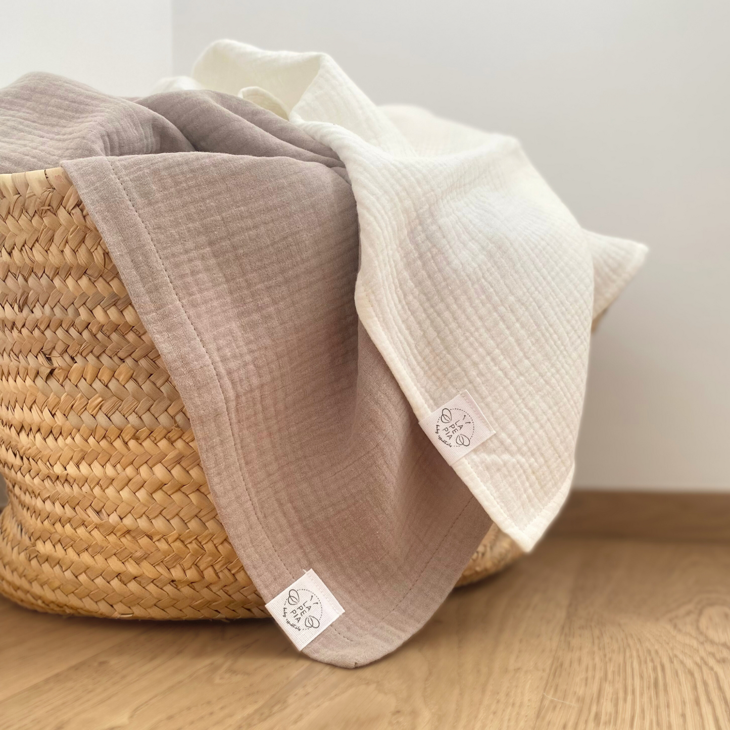 Copertine in mussola di cotone nei colori cream e beige appoggiate su di una cesta in paglia.