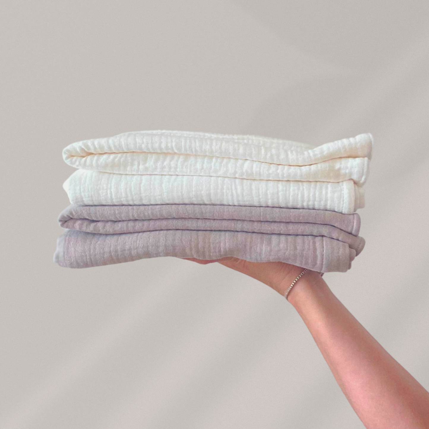 Copertine in mussola di cotone nei colori beige e cream, piegate e appoggiate sul palmo della mano.