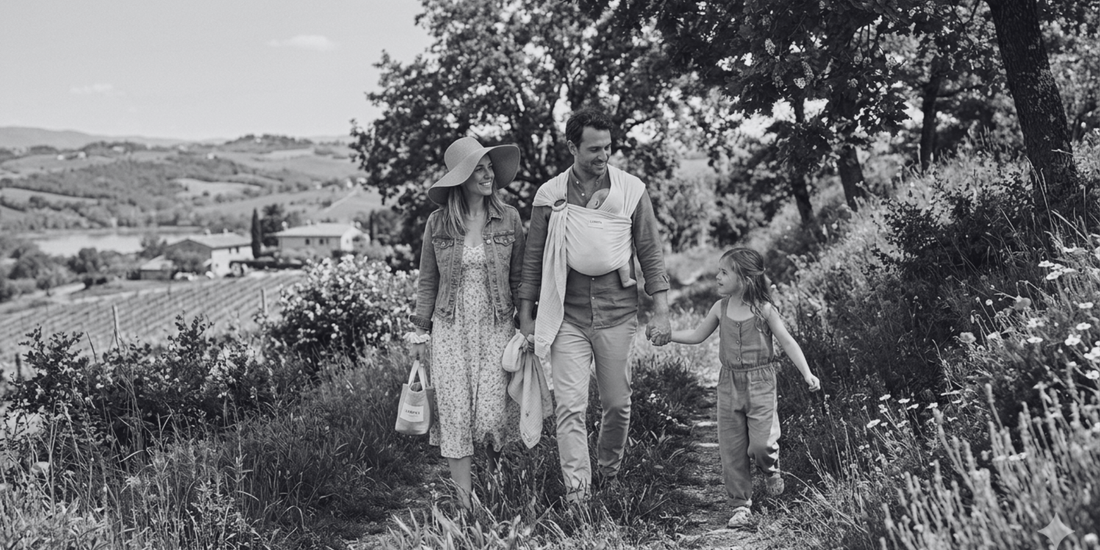 Una famiglia di quattro persone cammina serenamente lungo un sentiero sterrato in un paesaggio collinare lussureggiante e fiorito, sotto la calda luce del sole primaverile.