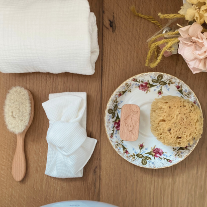 Ambientazione su piano in legno con spazzola in legno FSC e setole naturali, mussola color cream e spugna naturale con spazzolina per unghie appoggiati su piattino.