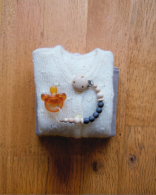 Flat lay di accessori per neonato con clip portaciuccio in legno di faggio, ciuccio anatomico in caucciù e maglione in maglia.