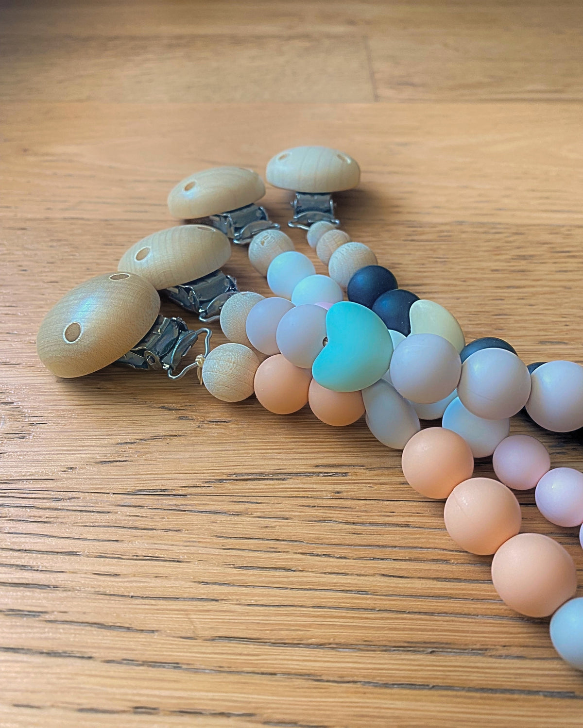Assortimento di catenelle portaciuccio in silicone alimentare e legno con perle colorate su tavolo in legno.