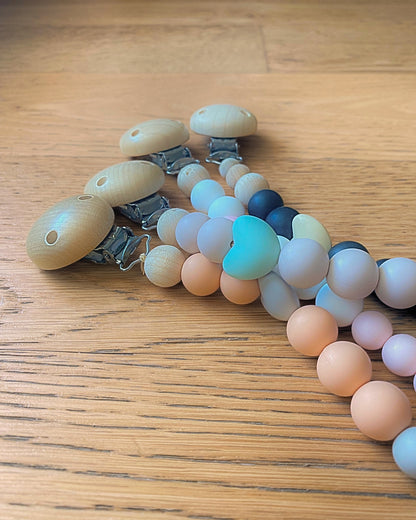 Assortimento di catenelle portaciuccio in silicone alimentare e legno con perle colorate su tavolo in legno.
