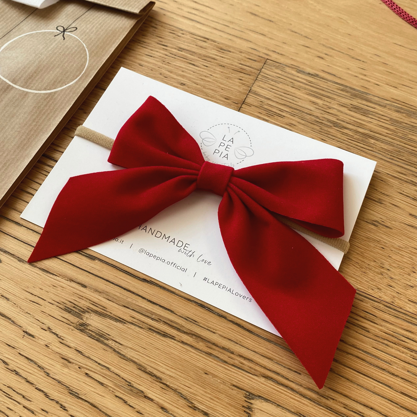 Fiocco per capelli in velluto rosso fotografato su cartolina e confezione sullo sfondo