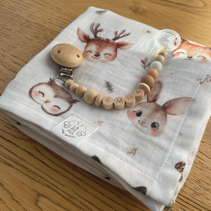 Mussola di cotone 70x70 variante Bosco Incantato ben piegata su un piano in legno, con sopra la catenella portaciuccio personalizzata LAPEPIA, accessori coordinati per neonati e bambini.